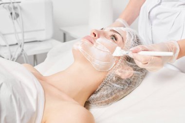 Güzellik uzmanı, güzellik salonlarında cilt bakımı için kadın yüzüne vitamin kremi maskesi, yaşlanma karşıtı kozmetik işlemler uyguluyor. Eldivenli kozmetik uzmanı maske takmak için kozmetik fırça tutuyor