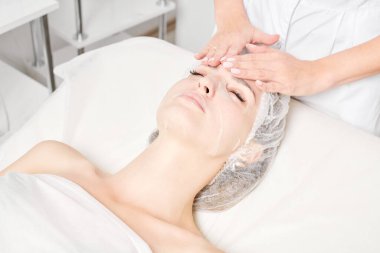 Güzellik uzmanı, güzellik salonundaki cilt bakımı için kadın yüzüne çarşaf maskesi, kırışıklık önleyici kozmetik prosedürler takıyor. Kozmetik uzmanı kadın yüzüne nemlendirici çarşaf maskesi uyguluyor.