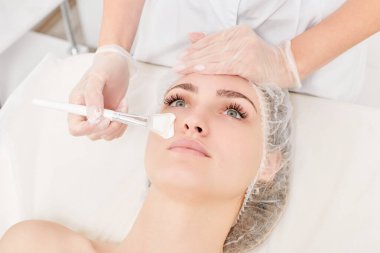Güzellik uzmanı, güzellik salonlarında cilt bakımı için kadın yüzüne vitamin kremi maskesi, yaşlanma karşıtı kozmetik işlemler uyguluyor. Eldivenli kozmetik uzmanı maske takmak için kozmetik fırça tutuyor