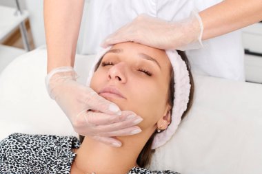 Eldivenli kozmetik uzmanı yenilenme, lenfatik drenaj, güzellik salonundaki krem cilt estetiği için kadın yüz masajı yapıyor. Güzellik uzmanı elleriyle cildini düzleştiriyor.