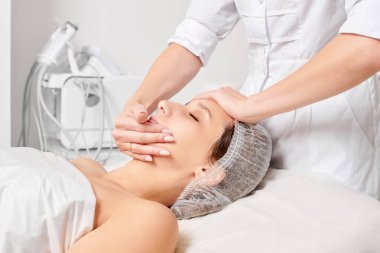 Güzellik salonunda kozmetik uzmanı cildine nemlendirici krem maskeyi sürüp gençleştirme, yaşlanmayı önleyici kozmetik prosedür uyguluyor. Güzellik uzmanı elleri kadın derisi için yüz kremi sürüyor.