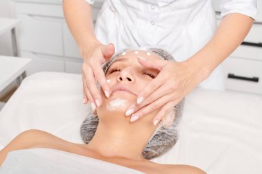Güzellik uzmanları, güzellik salonlarında gençleştirme için nemlendirici krem maskeyi kadın yüzüne sürerler, yaşlanma karşıtı kozmetik işlemler yaparlar. Kozmetik uzmanı kadın derisi için yüz kremi uyguluyor.