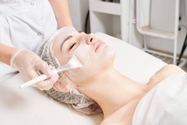 Güzellik uzmanı, güzellik salonlarında cilt bakımı için kadın yüzüne vitamin kremi maskesi, yaşlanma karşıtı kozmetik işlemler uyguluyor. Eldivenli kozmetik uzmanı maske takmak için kozmetik fırça tutuyor