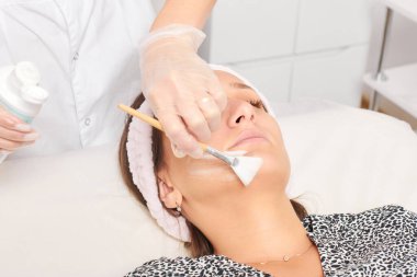 Güzellik uzmanı, güzellik salonundaki kozmetik krem maskesini cilt tazeleme, nemlendirici kozmetik işlemler için kadın yüzüne uyguluyor. Eldivenli kozmetik uzmanı fırçayla kozmetik krem maskesi takıyor.