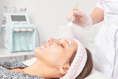 Kozmetik uzmanı, güzellik salonundaki kozmetik bakım ürünlerini nemlendirmek için kadın yüzüne estetik krem takıyor. Eldivenli güzellik uzmanı fırçayla kozmetik krem maskesi takıyor.