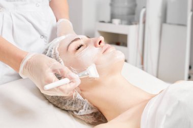 Güzellik uzmanı, güzellik salonundaki yaşlanma karşıtı kozmetik işlemler için cildini yenilemek için kadın yüzüne kozmetik krem maskesi sürüyor. Eldivenleri teslim eden kozmetik uzmanı maske takmak için kozmetik fırça tutuyor.