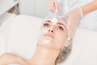 Güzellik uzmanı, güzellik salonundaki yaşlanma karşıtı kozmetik işlemler için cildini yenilemek için kadın yüzüne kozmetik krem maskesi sürüyor. Eldivenleri teslim eden kozmetik uzmanı maske takmak için kozmetik fırça tutuyor.