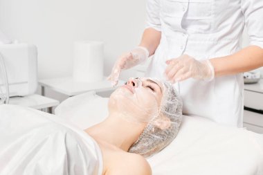 Güzellik uzmanı güzellik salonundaki nemlendirici cilt bakım için kadın yüzüne krem maskesi takıyor. Kozmetik uzmanı kadın yüzüne krem sürerken eldiven verdi.
