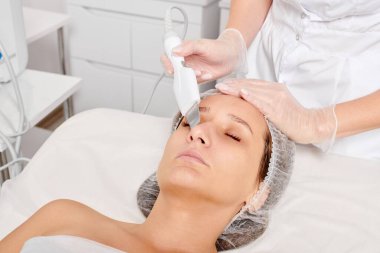 Kozmetik uzmanı, güzellik salonundaki yaşlanma karşıtı kozmetik prosedürleri, cilt bakımını yenilemek için yüz ultrasoniği yapıyor. Güzellik uzmanı yüz bakımını ultrason fırçasıyla yapıyor.