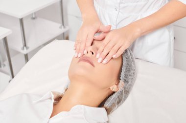 Güzellik uzmanı, güzellik salonunda gençleştirme için nemlendirici krem sürdükten sonra cildine masaj yapıyor, yaşlanma önleyici kozmetik prosedürler uyguluyor. Kozmetik uzmanı rahatlamak için kadın yüzüne masaj yapıyor.