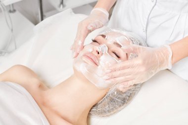Kozmetik uzmanı, güzellik salonundaki yaşlanma karşıtı kozmetik prosedürleri ve cilt bakımı için kadın yüzüne krem masajı yapıyor. Eldivenli güzellik uzmanı elleri kadın yüzüne krem sürüyor.