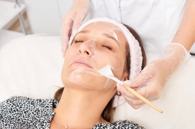 Kozmetik uzmanı, güzellik salonundaki kozmetik bakım ürünlerini nemlendirmek için kadın yüzüne estetik krem takıyor. Eldivenli güzellik uzmanı fırçayla kozmetik krem maskesi takıyor.
