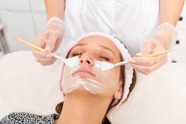 Güzellik uzmanı, güzellik salonunda estetik krem maskesini cilt tazeleme, nemlendirici kozmetik işlemleri için kadın yüzüne uyguluyor. Eldivenli kozmetik uzmanı iki fırçayla kozmetik krem maskesi takıyor.