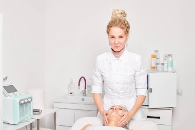 Güzellik salonunda kozmetik uzmanı cildine nemlendirici krem maskeyi sürüp gençleştirme, yaşlanmayı önleyici kozmetik prosedür uyguluyor. Güzellik uzmanı elleri kadın derisi için yüz kremi sürüyor.