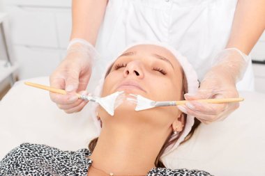 Güzellik uzmanı, güzellik salonunda estetik krem maskesini cilt tazeleme, nemlendirici kozmetik işlemleri için kadın yüzüne uyguluyor. Eldivenli kozmetik uzmanı iki fırçayla kozmetik krem maskesi takıyor.