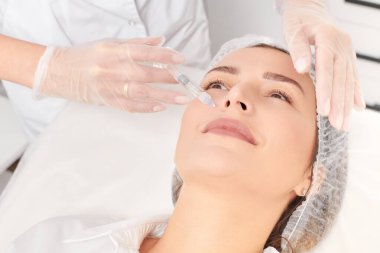 Kozmetik uzmanı kadın tenine gençleştirme enjeksiyonu yapıyor güzellik salonundaki yaşlanma karşıtı kozmetik prosedürü. Eldivenli güzellik uzmanı yüz asidi enjeksiyonu tedavisi yapıyor.