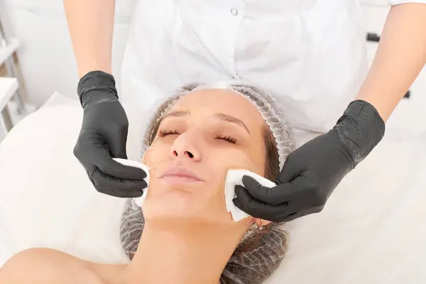 Beautician removing facial cream mask from woman face skin with cosmetic wipes, rejuvenation skincare cosmetic procedure in beauty spa salon. Cosmetologist hands holds napkins for remove face mask