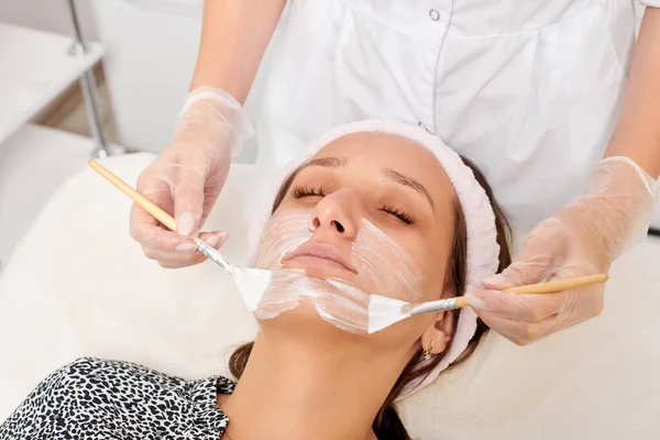 Kozmetik uzmanı, güzellik salonunda cildini yenilemek için kadın yüzüne kozmetik krem maskesi uyguluyor. Eldivenli güzellik uzmanı iki fırçalı kozmetik krem maskesi takıyor.