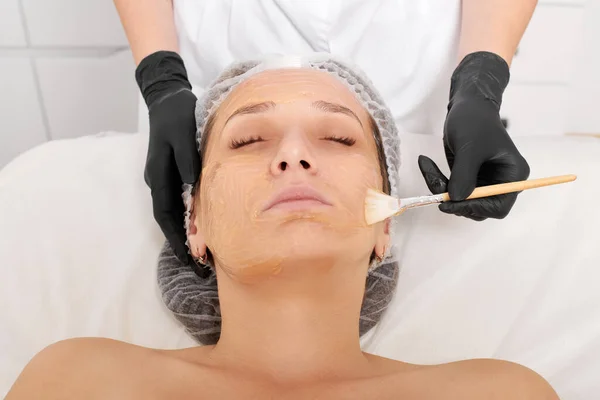 Cosmetologist applies honey mask on woman face for moisturize face skin, anti aging cosmetic procedure in beauty spa salon. Beautician in black gloves holds cosmetic brush for applying honey mask