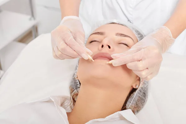 Beautician applies facial oil capsules on woman face for rejuvenation, anti aging cosmetic procedure in beauty spa salon. Cosmetologist hands in gloves holds oil capsules for nourishment