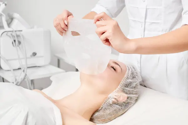 Cosmetologist applying sheet mask on woman face for rehydrate face skin, revitalize cosmetic procedure in beauty spa salon. Beautician applying detox sheet mask on female face