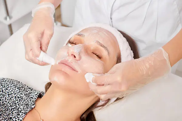 Cosmetologist removing facial cream mask from woman face skin with cosmetic wipes, rejuvenation skincare cosmetic procedure in beauty spa salon. Beautician removing moisturized cream with napkins