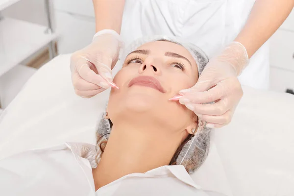Beautician applies facial oil capsules on woman face for rejuvenation, anti aging cosmetic procedure in beauty spa salon. Cosmetologist hands in gloves holds oil capsules for nourishment