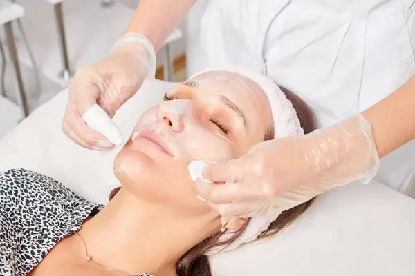 Beautician removing facial cream mask from woman face skin with cosmetic wipes, rejuvenation skincare cosmetic procedure in beauty spa salon. Cosmetologist removing moisturized cream with napkins