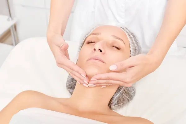 Cosmetologist massages woman face skin for rejuvenation, anti aging skincare cosmetic procedure in beauty spa salon. Beautician making facial massage with circular movements for beautiful female face