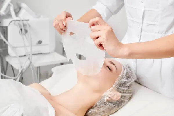 Beautician applies sheet mask on woman face for rehydrate face skin, anti aging cosmetic procedure in beauty spa salon. Cosmetologist applying moisturizing sheet mask on female face