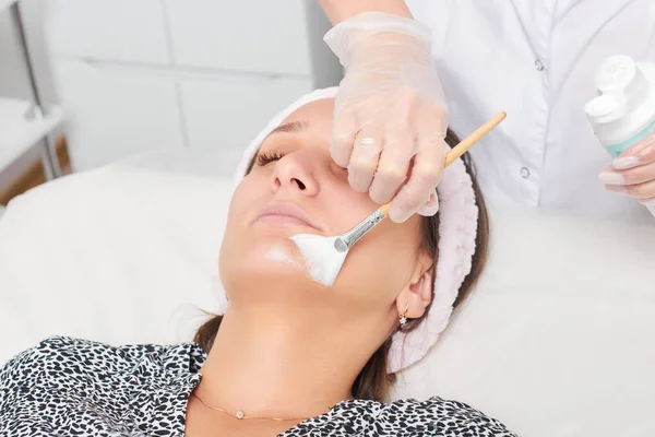 Kozmetik uzmanı, güzellik salonundaki kozmetik bakım ürünlerini nemlendirmek için kadın yüzüne estetik krem takıyor. Eldivenli güzellik uzmanı fırçayla kozmetik krem maskesi takıyor.
