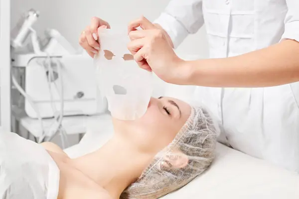 Beautician applies sheet mask on woman face for rehydrate face skin, anti aging cosmetic procedure in beauty spa salon. Cosmetologist applying moisturizing sheet mask on female face