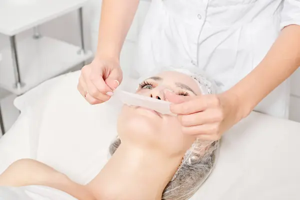 Beautician removing sheet mask on woman face for rehydrate face skin, anti aging cosmetic procedure in beauty spa salon. Cosmetologist removing moisturizing sheet mask on female face