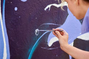 Kadın ressam, açık hava sokak sergisi için tuval üzerine boya fırçasıyla resim çiziyor, kadın ressamın arka yüzünü kapatıyor, fırça darbelerini dış mekâna hayat aşılayan tuvale uyguluyor.