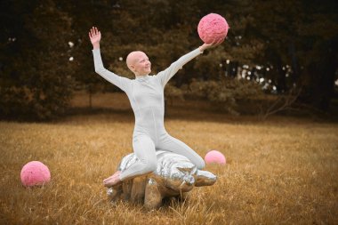 Beyaz giysili tüysüz genç kız tardigrad figürünün üzerinde oturuyor ve sonbahar çim parkında pembe topu elinde tutuyor. Kel genç kızın sembolik elementlerle nişanlandığı gerçeküstü bir sahne.