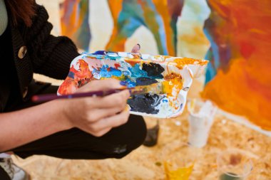 Kadın ressam el fırçasını açık hava sergisi için canlı resim paletine daldırıyor, elinde fırça tutan kadın ressamın görüntüsünü kapatıyor.