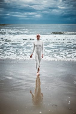 Beyaz fütüristik takım elbiseli tüysüz kız kumlu sahildeki soğuk ve huzursuz denizden çıkıyor. Kel kadın sanatçının hayatın zorluklarını aşma ve kendini kabullenme metaforik performansı.