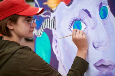 Kırmızı şapkalı kadın ressam, açık hava sokak sergisi için tuval üzerine fırçayla gerçeküstü resim çiziyor, kadın ressamın yan görüntüsünü kapatıyor, tuvale fırça darbeleri uyguluyor, sanat yaratıcılığı senfonisi.