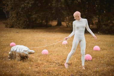 Saçsız genç bir kızın portresi beyaz kumaş içinde tardigrad oyuncağı sonbahar parkında, kel genç bir kızla gerçeküstü bir sahnede hayat ve sanatın iç içe geçişini yansıtıyor.