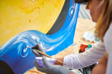 Siyah şapkalı kadın ressam, açık hava sergisi için tuval üzerine fırçayla resim çiziyor, kadın ressamın yan görüntüsünü kapatıyor fırça darbelerini tuvale uyguluyor, sanat yaratıcılığı senfonisi.