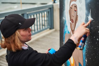 Siyah şapkalı kadın ressam, boya kutusuyla resim yapıyor. Açık hava sokak sergisinde tuvale püskürtüyor. Kadın sanatçının yan görüntüsü.