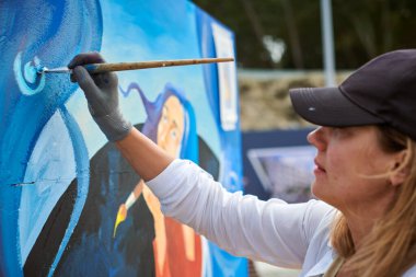 Siyah şapkalı kadın ressam, açık hava sergisi için tuval üzerine fırçayla resim çiziyor, kadın ressamın yan görüntüsünü kapatıyor fırça darbelerini tuvale uyguluyor, sanat yaratıcılığı senfonisi.