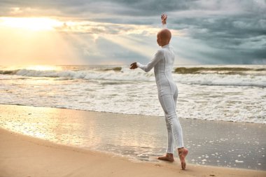 Young hairless ballerina with alopecia in white futuristic suit dancing on seashore at sunset sea, metaphoric surreal performance of bald pretty teenage girl exudes confidence, hope and unique beauty clipart