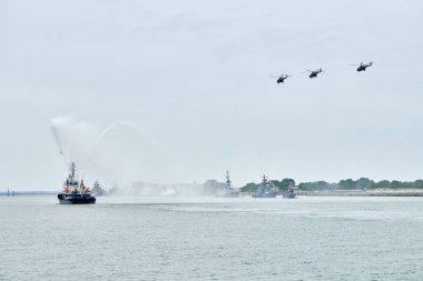 Rus deniz kuvvetleri boyunca yelken açan itfaiye teknesi kıyı şeridi boyunca askeri helikopterlerle savaş gemilerini geçit töreni, askeri gemilerin oluşumu denizcilik geleneği, Rus deniz gücünün deniz manzarası.