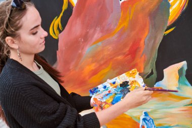 Çekici kadın ressam, açık hava sokak sergisi için boya fırçasıyla resim çiziyor, açık hava sanat alanına hayat veren kadın ressamın yan görüntüsünü kapatıyor.