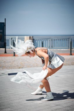 Beyaz ipek eşarpla dans eden, uzay gümüş renkli mini etekli genç seksi kız, deniz kenarında dans eden kadın, heyecan verici bir açık hava gösterisi yaratıyor.