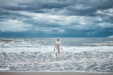 Beyaz fütüristik takım elbiseli tüysüz kız kumlu sahildeki soğuk ve huzursuz denizden çıkıyor. Kel kadın sanatçının hayatın zorluklarını aşma ve kendini kabullenme metaforik performansı.
