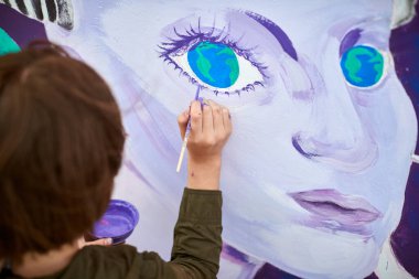 Kadın ressam, açık hava sokak sergisi için tuvale boya fırçasıyla resim çiziyor, kadın ressamın üst görüşünü kapatıyor, fırça darbelerini tuvale uyguluyor, sanat yaratıcılığı senfonisi