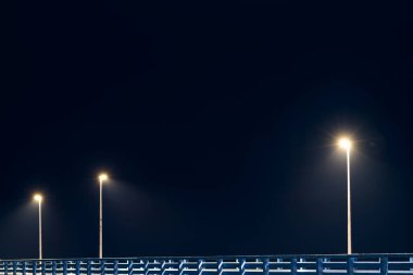 Street lampposts casts dim cold blue glow along shore pier standing against pitch black canvas of night sky paints poetic picture of pier in stillness of night, serenity of solitary moments clipart