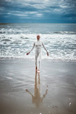 Beyaz fütüristik takım elbiseli tüysüz kız kumlu sahildeki soğuk ve huzursuz denizden çıkıyor. Kel kadın sanatçının hayatın zorluklarını aşma ve kendini kabullenme metaforik performansı.